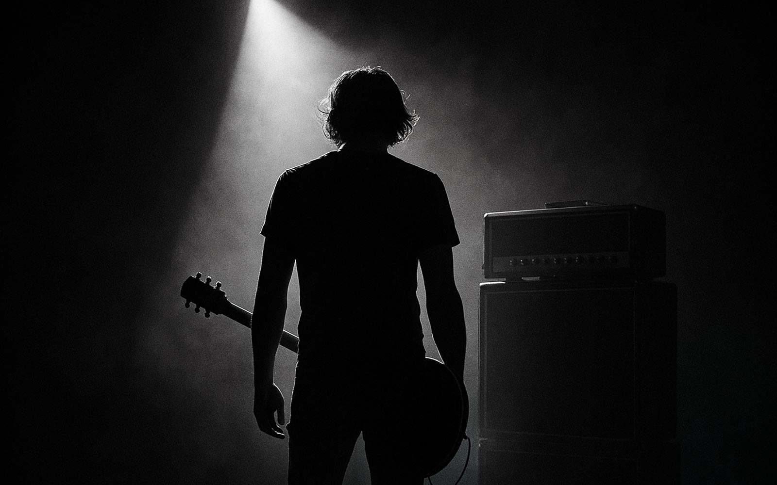 Black-and-white photo of a solitary guitarist under a spotlight in front of a half-stack—focused practice, discipline, intent.