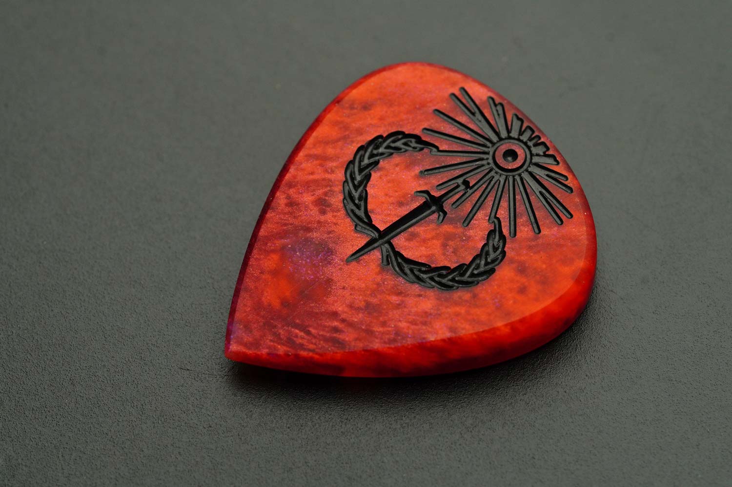 Sol Invictus red resin guitar pick shown flat with black sun, sword, and laurel engraving