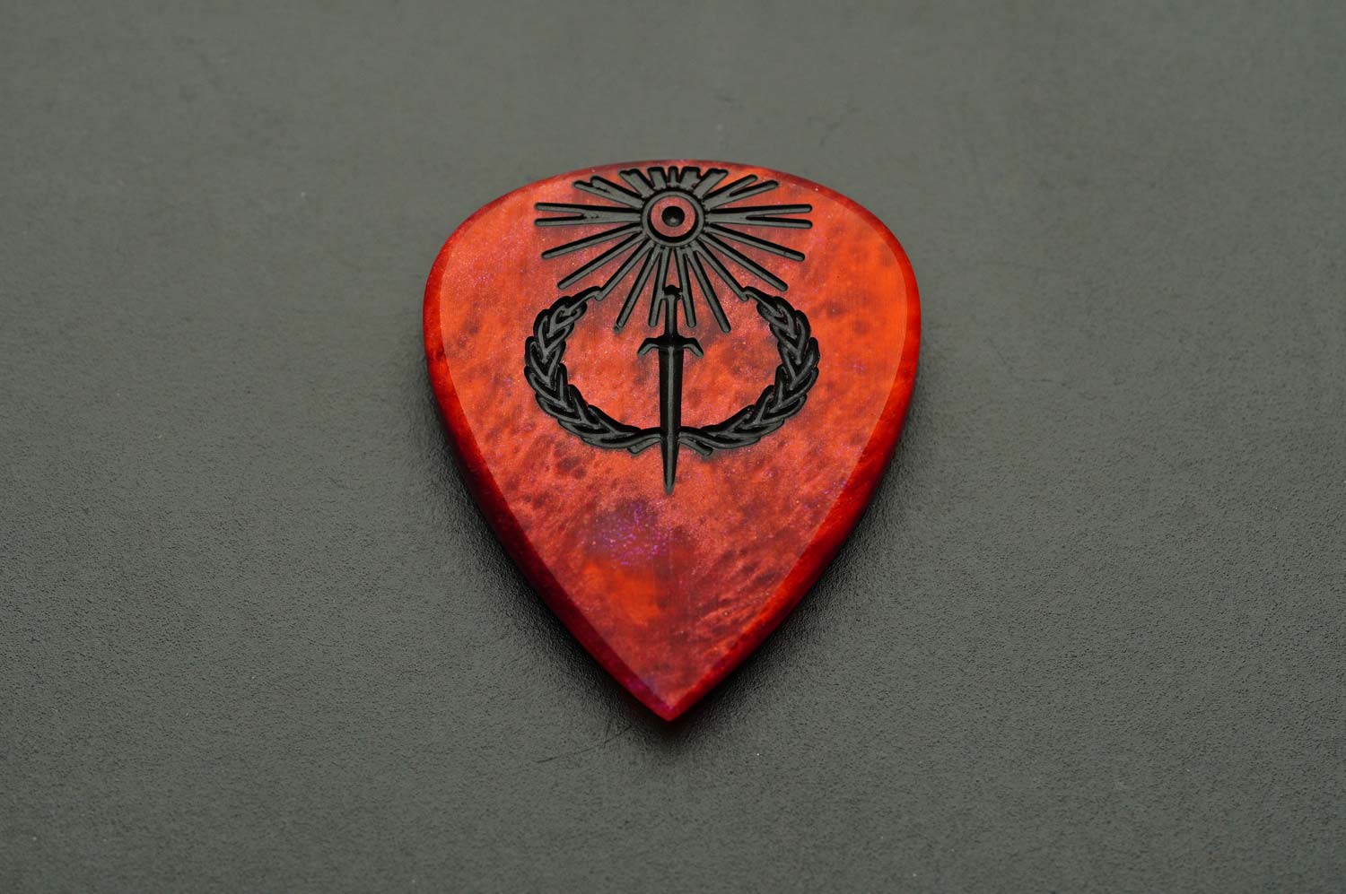 Sol Invictus red resin guitar pick shown flat with black sun, sword, and laurel engraving